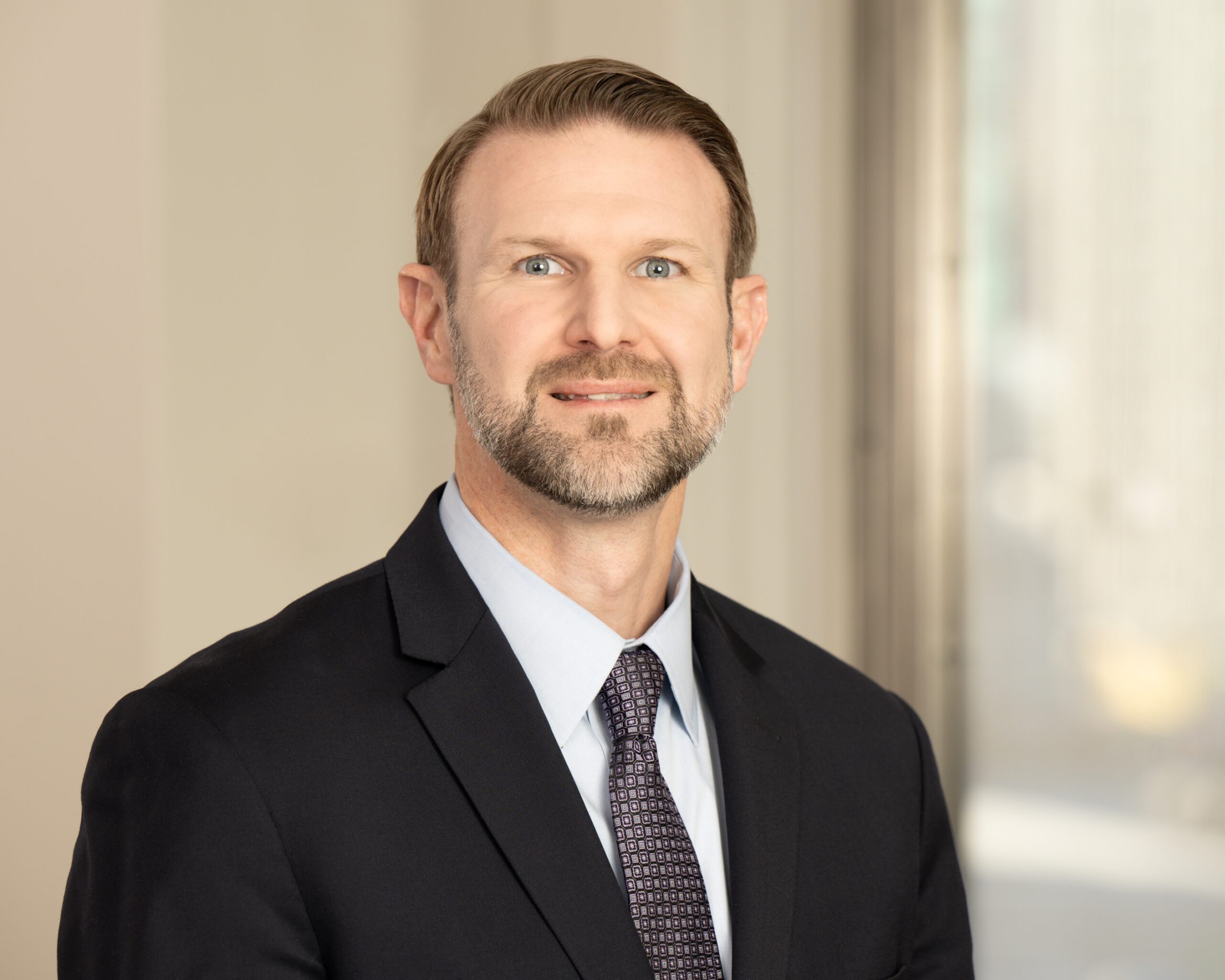 A man with brown hair, a close-trimmed brown and grey beard and wearing dark-grey suit with a light blue shirt and dark purple and white patterned tie is seen in an office smiling toward the camera
