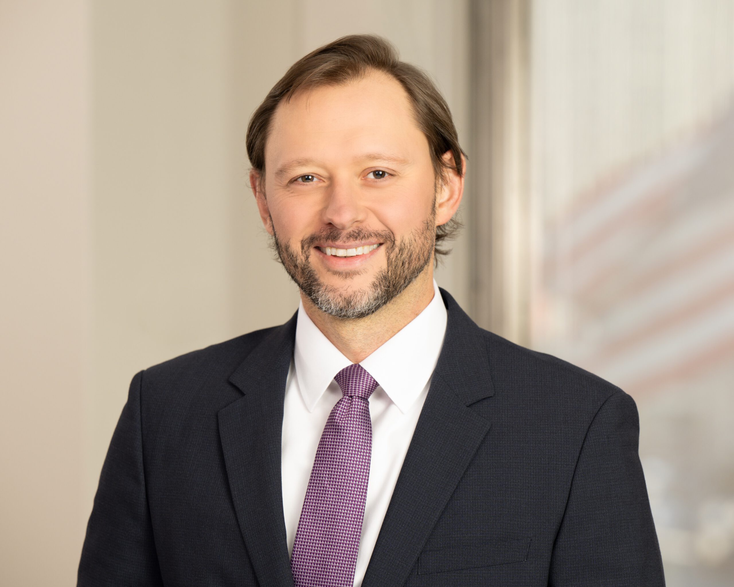 A man with dark brown hair and a close-trimmed, brown and grey beard, wearing a dark, blue and grey patterned suit, white-collared shirt, and a purple-and-white patterned tie is seen in an office setting smiling toward the camera