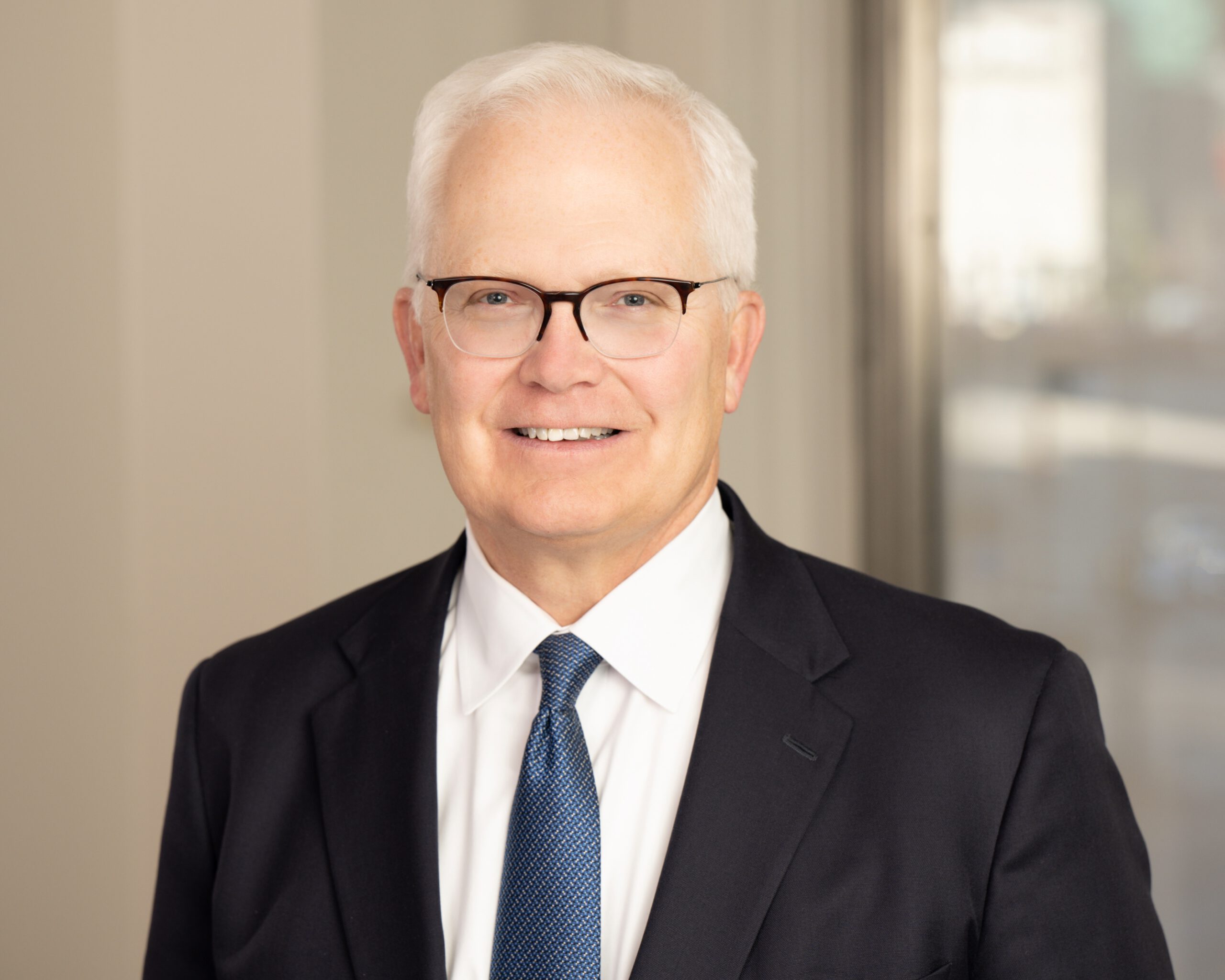 A man with short, light-grey hair wearing black, partially frameless glasses and a dark-grey suit with a white shirt and blue, patterned tie is seen in an office conference room smiling toward the camera